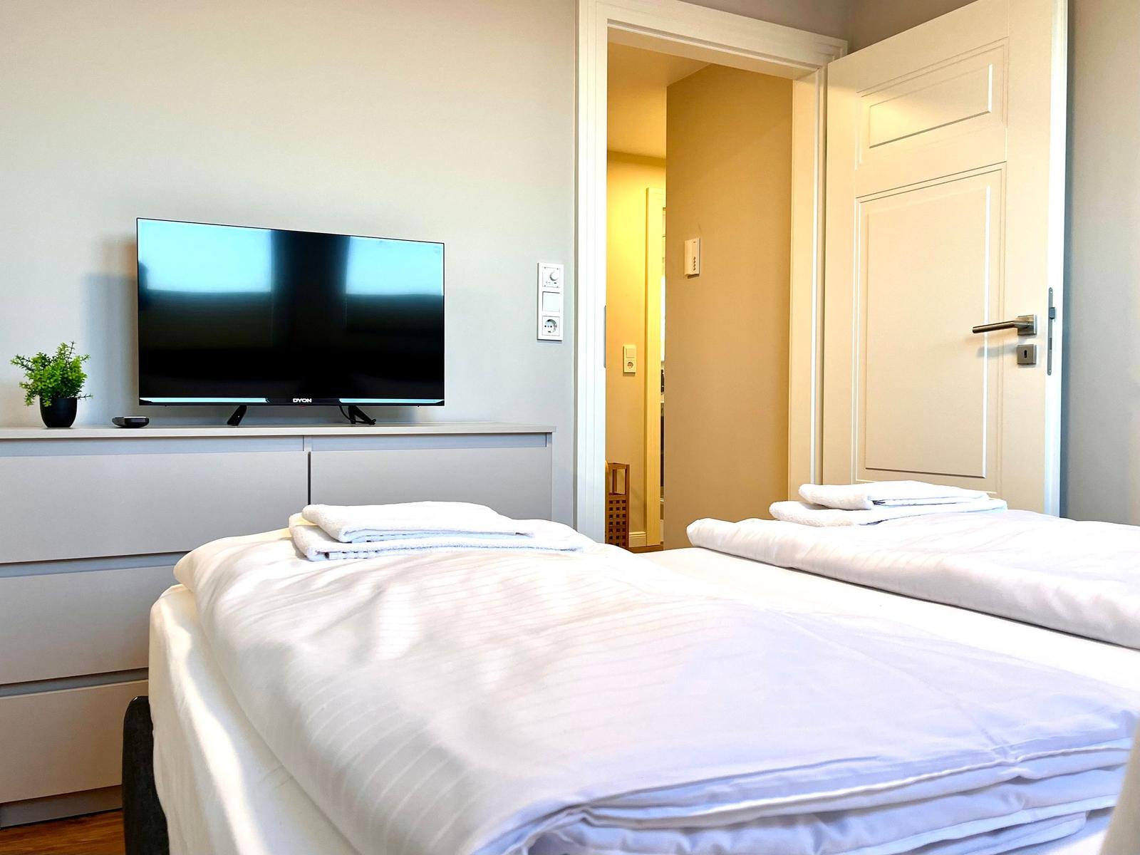 Zwei Betten mit weißen Laken, Fernseher und Tür zum Flur.
