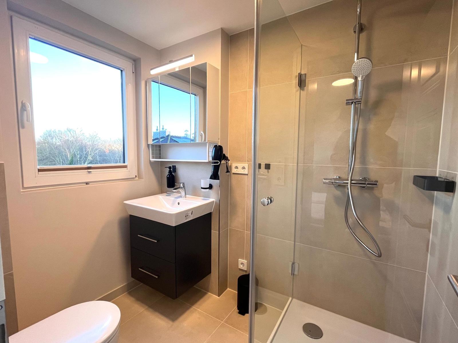 Badezimmer mit Dusche, Waschbecken und Fenster. Toilette und Spiegelschrank sichtbar.