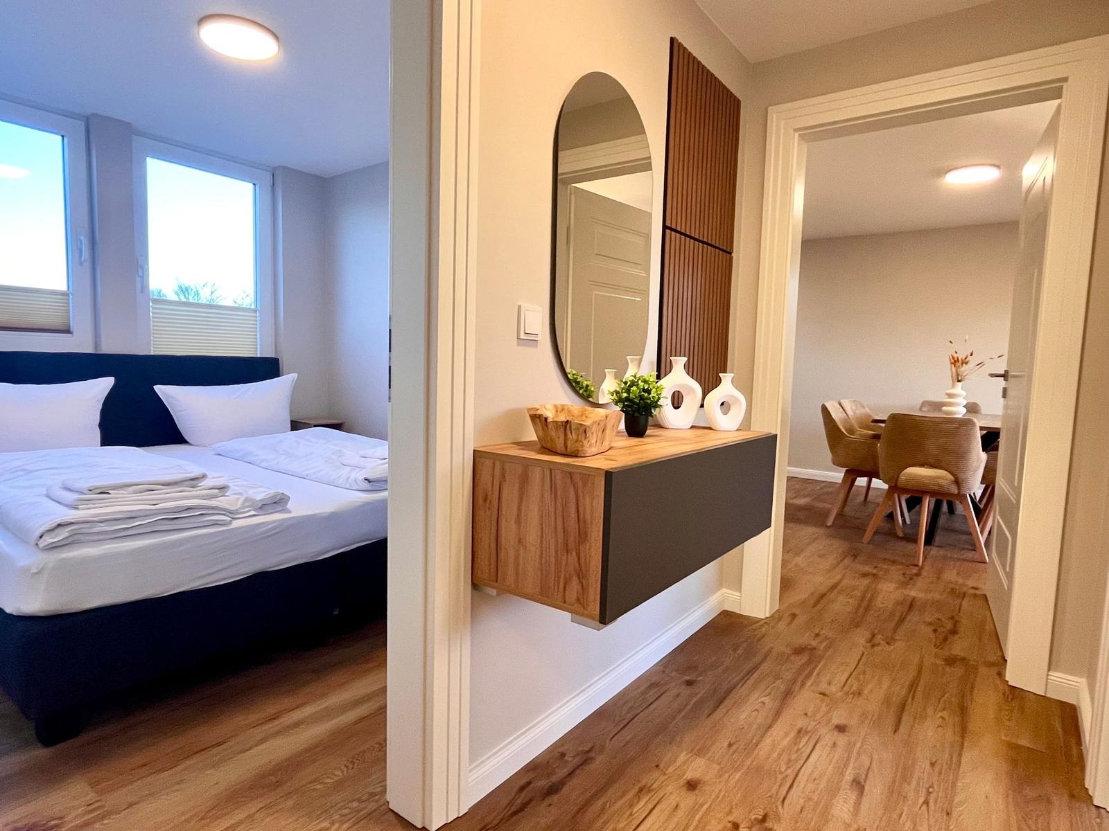 Schlafzimmer mit Bett und Fenstern. Flur mit Wandregal und Spiegel. Esszimmer mit Tisch und Stühlen.