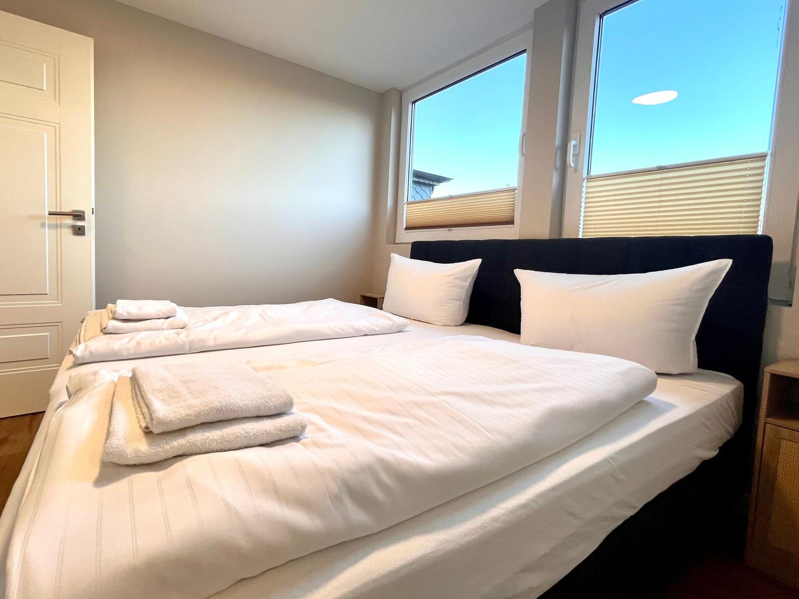 Schlafzimmer mit Doppelbett, weißen Laken und großen Fenstern.