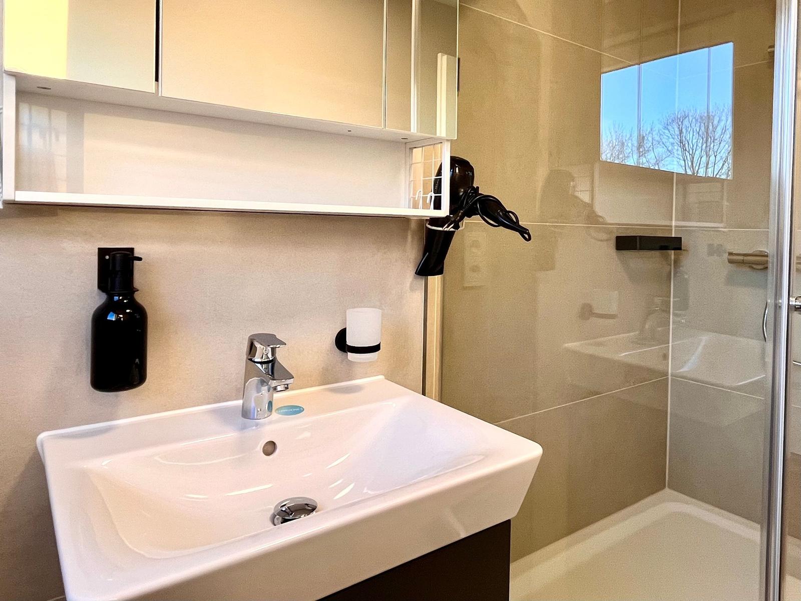 Bad mit Waschbecken, Spiegel und Dusche. Haartrockner und Seifenspender vorhanden.