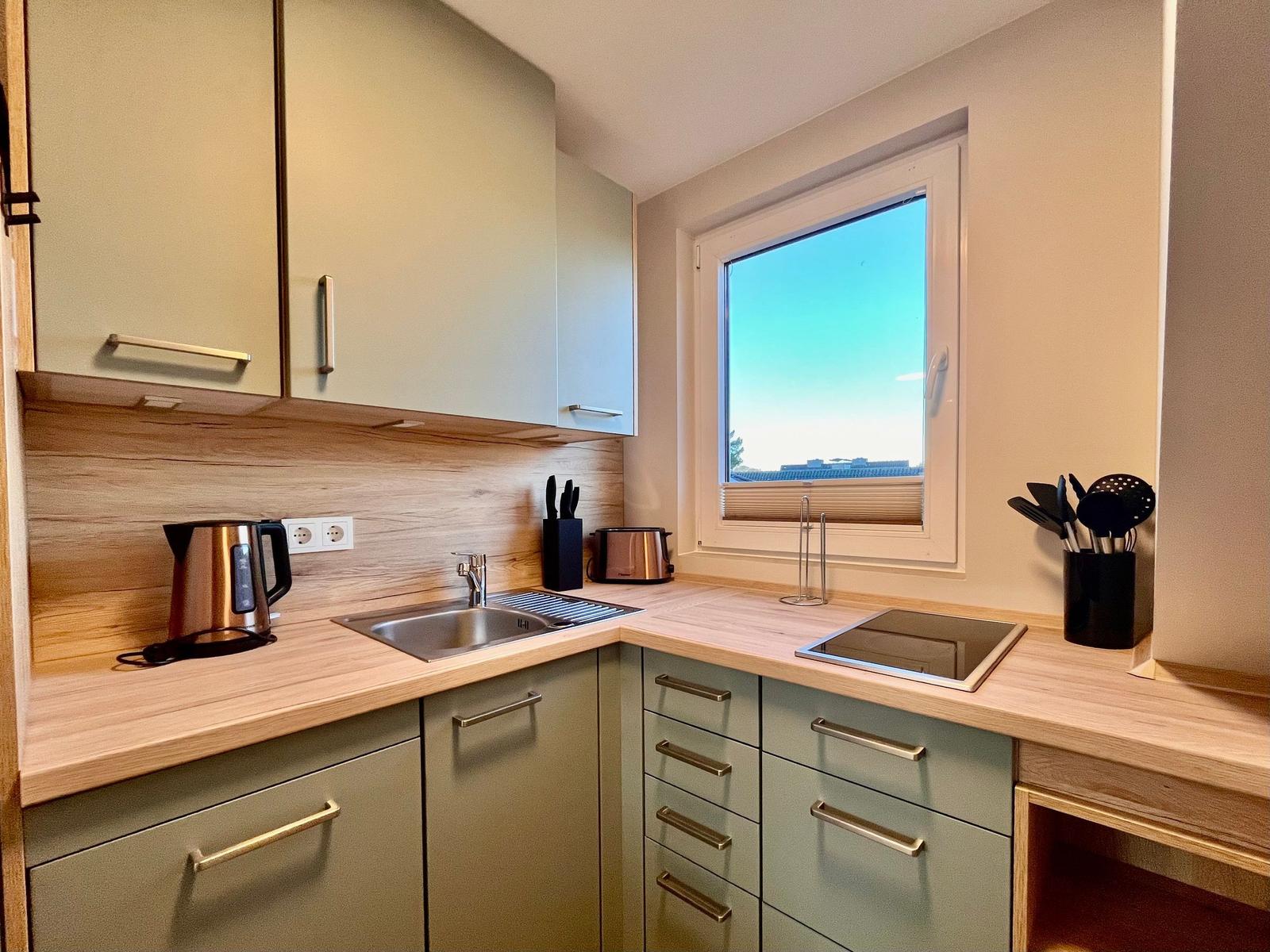 Küche mit Arbeitsfläche, Spüle, Herd und Fenster. Geschirr und Küchenutensilien sind vorhanden.