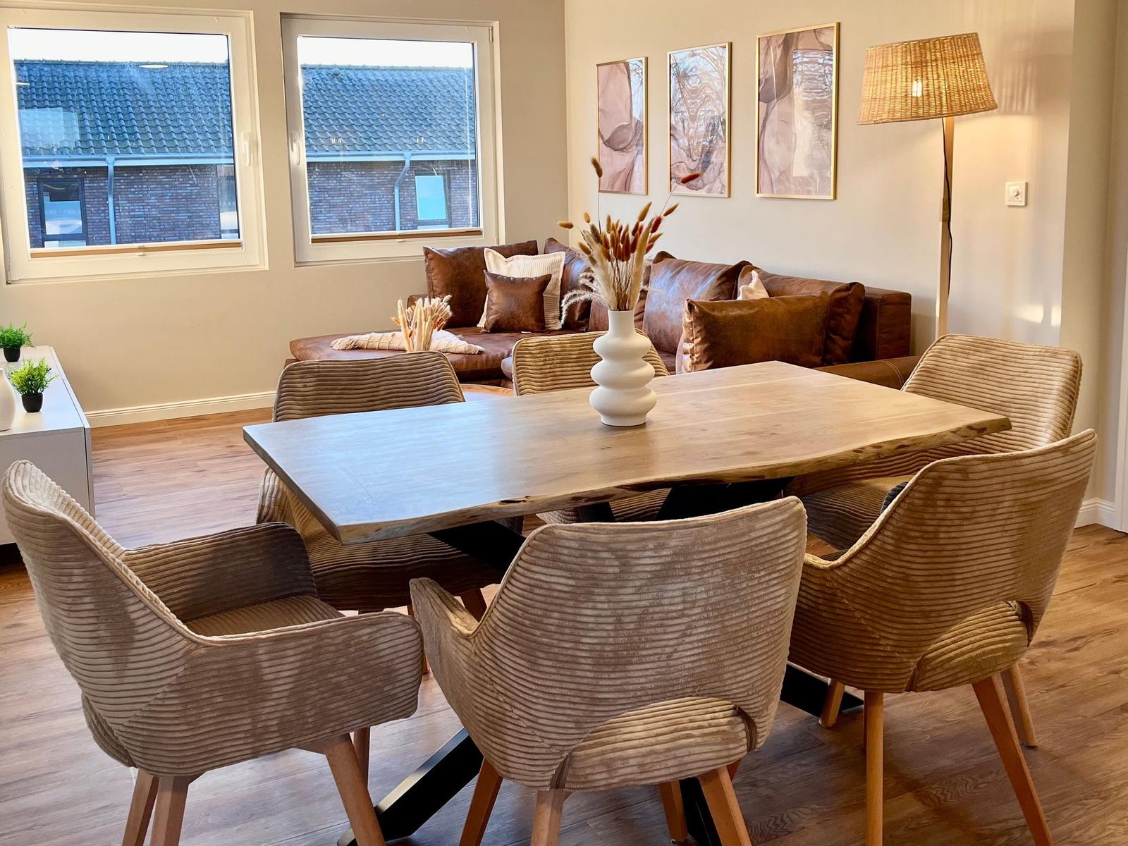 Essbereich mit Holztisch und sechs Stühlen. Im Hintergrund ein Sofa und Fenster.