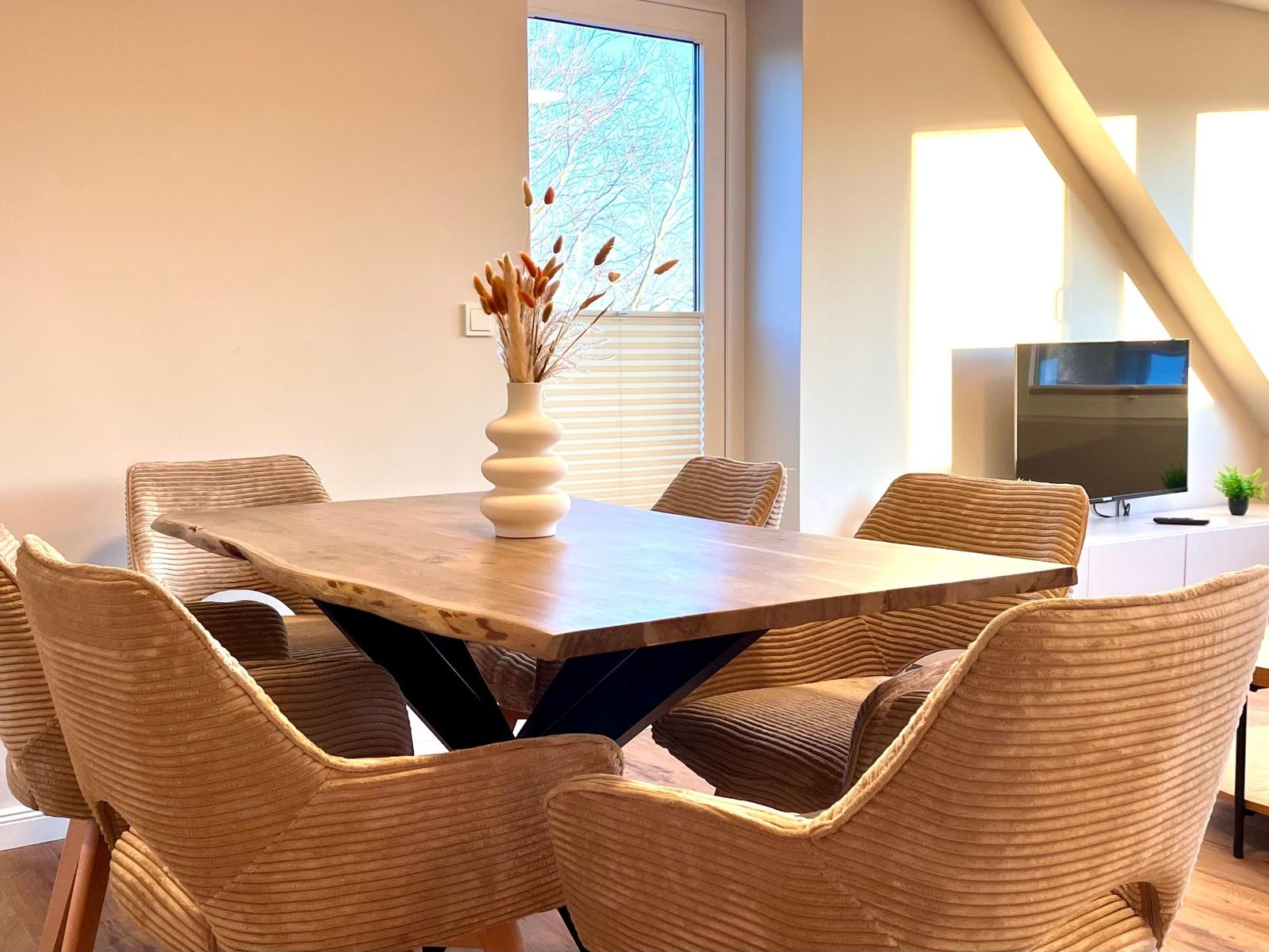 Esszimmer mit Holztisch, sechs Stühlen und Fenster. Fernseher im Hintergrund.
