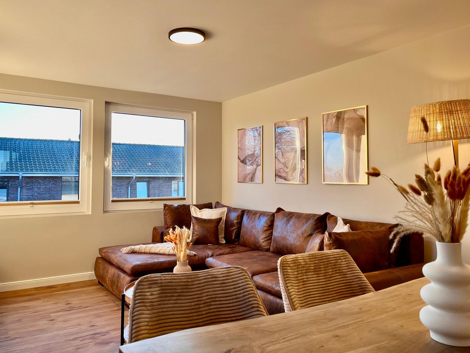 Wohnzimmer mit braunem Sofa, zwei Fenstern und drei Wandbildern.