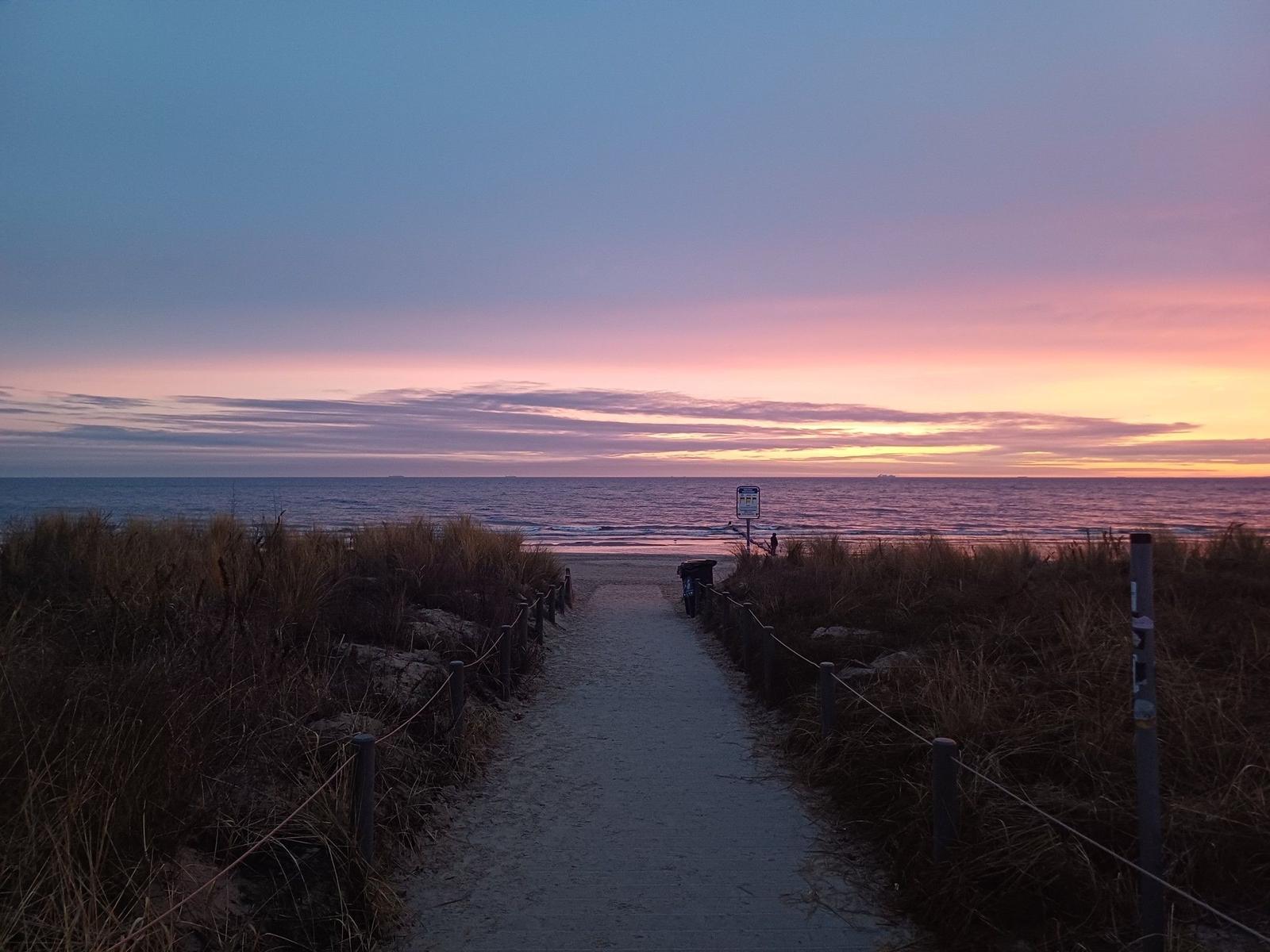 Sandweg führt zum Strand bei Sonnenuntergang mit rosa und blauem Himmel.