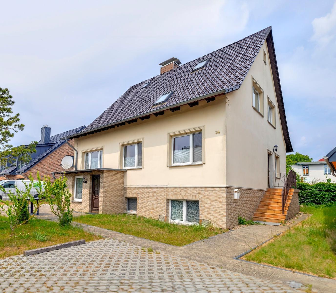 Ein zweistöckiges Haus mit braunem Dach und hellen Wänden. Eingangstreppe und gepflasterte Zufahrt.