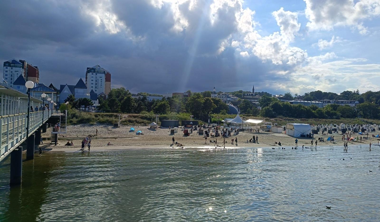 Strand mit Steg, Badegästen und Hotels im Hintergrund unter bewölktem Himmel.