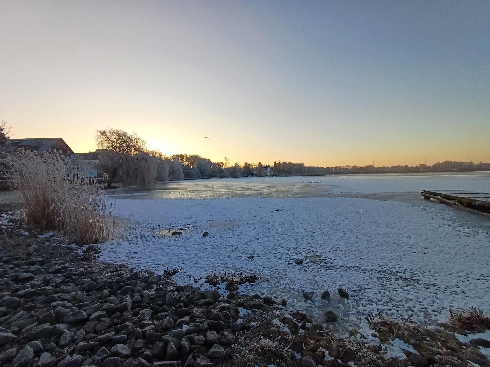 Eisiger See bei Sonnenuntergang mit schneebedeckten Ufern und Bäumen.