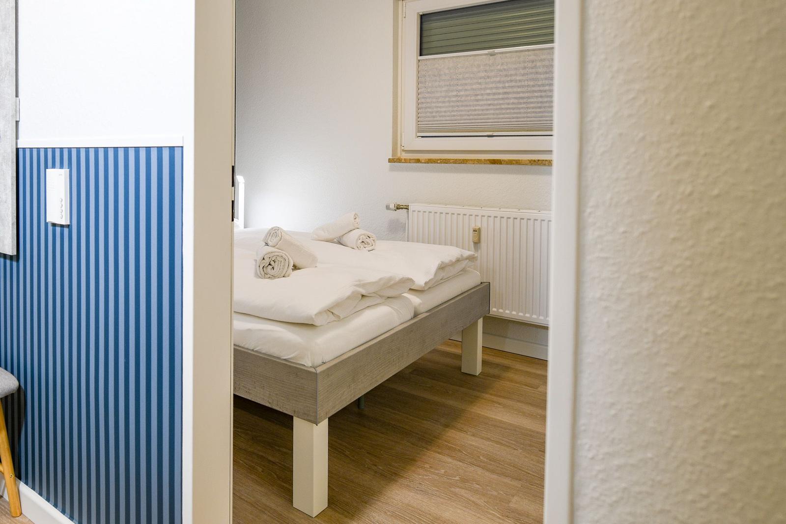 Schlafzimmer mit Bett, weißen Laken und Fenster mit Jalousie.