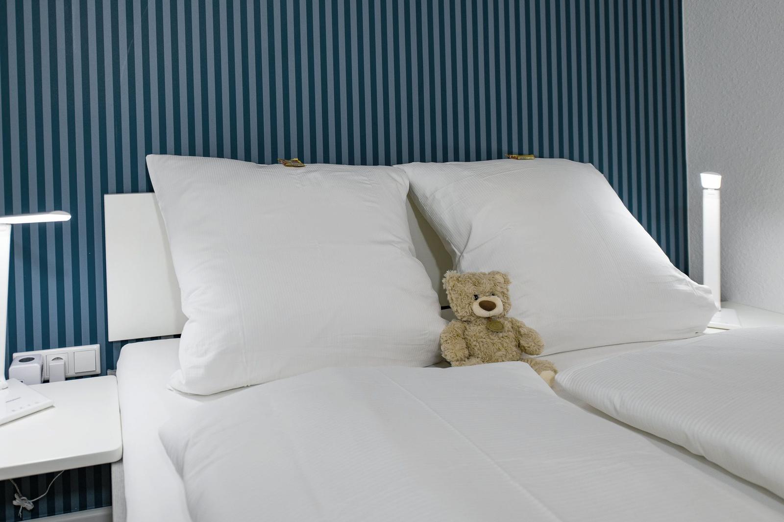 Doppelbett mit weißen Kissen und einem Teddybären. Streifenwand und Nachttisch.