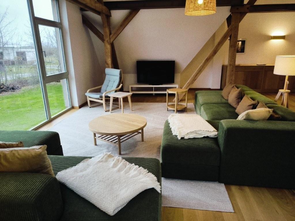 Wohnzimmer mit grüner Sofa, Holztisch und Fernseher. Große Fenster und Holzbalken.