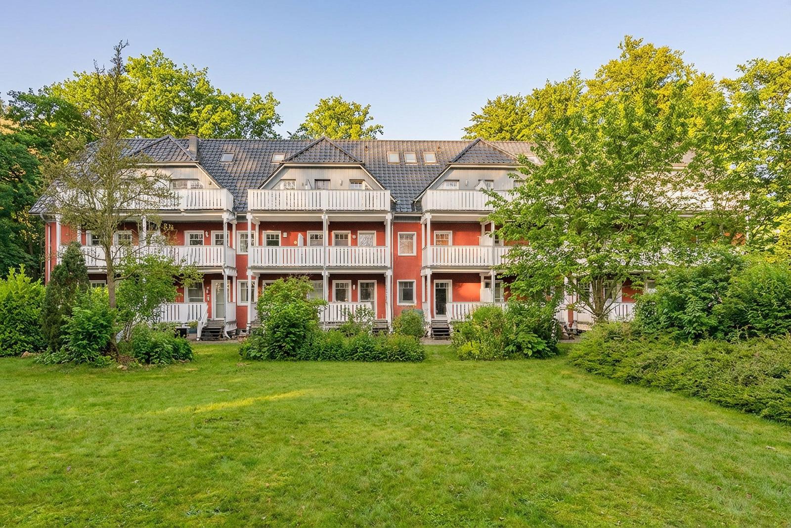 Rotes Mehrfamilienhaus mit weißen Balkonen und grünem Garten vor Bäumen.