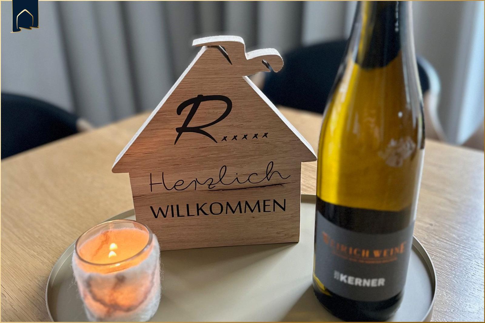 Willkommensschild, Kerze und Weinflasche auf Tisch.