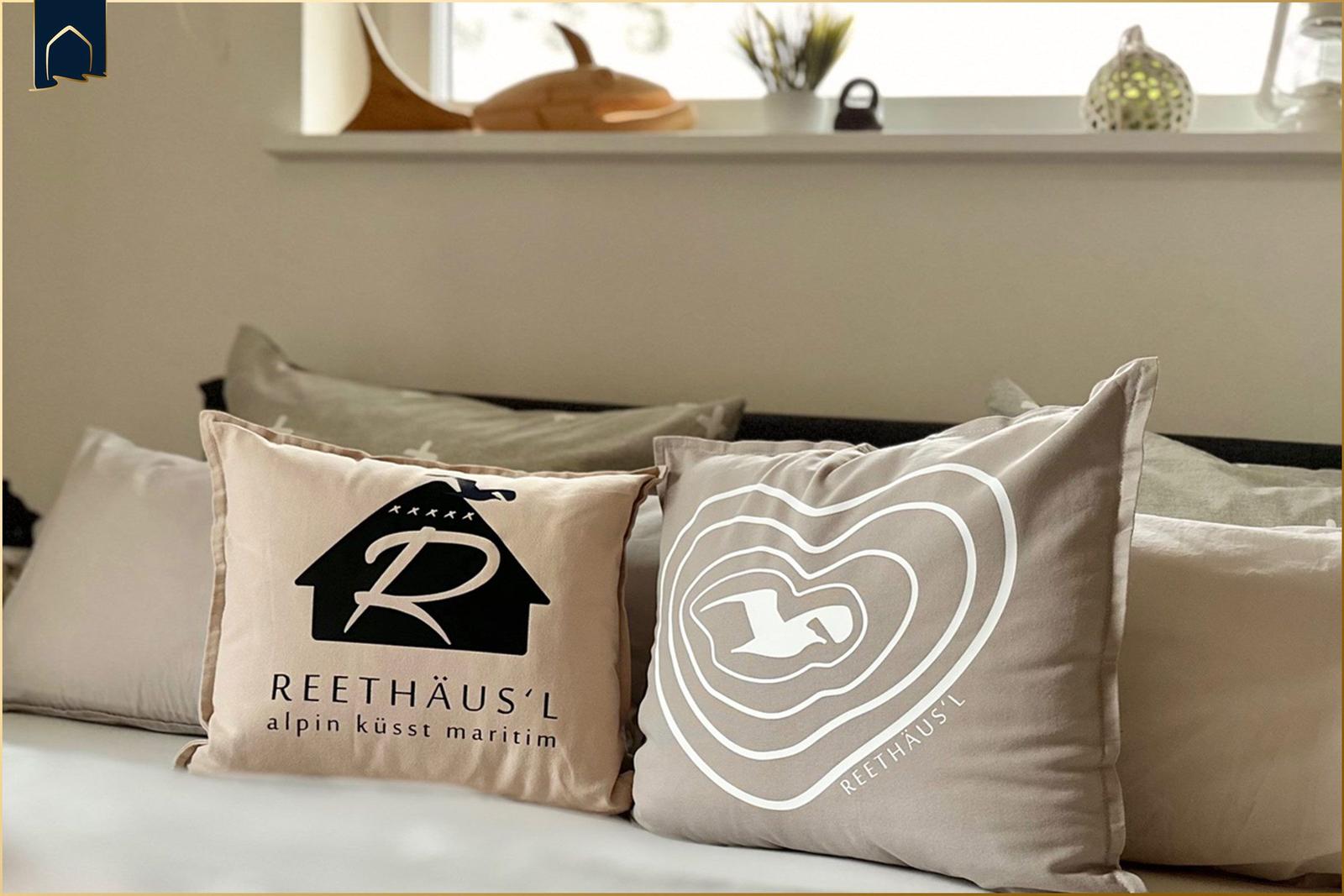 Zwei Kissen mit Logo auf Bett, Fensterbank mit Dekorationsgegenständen im Hintergrund.