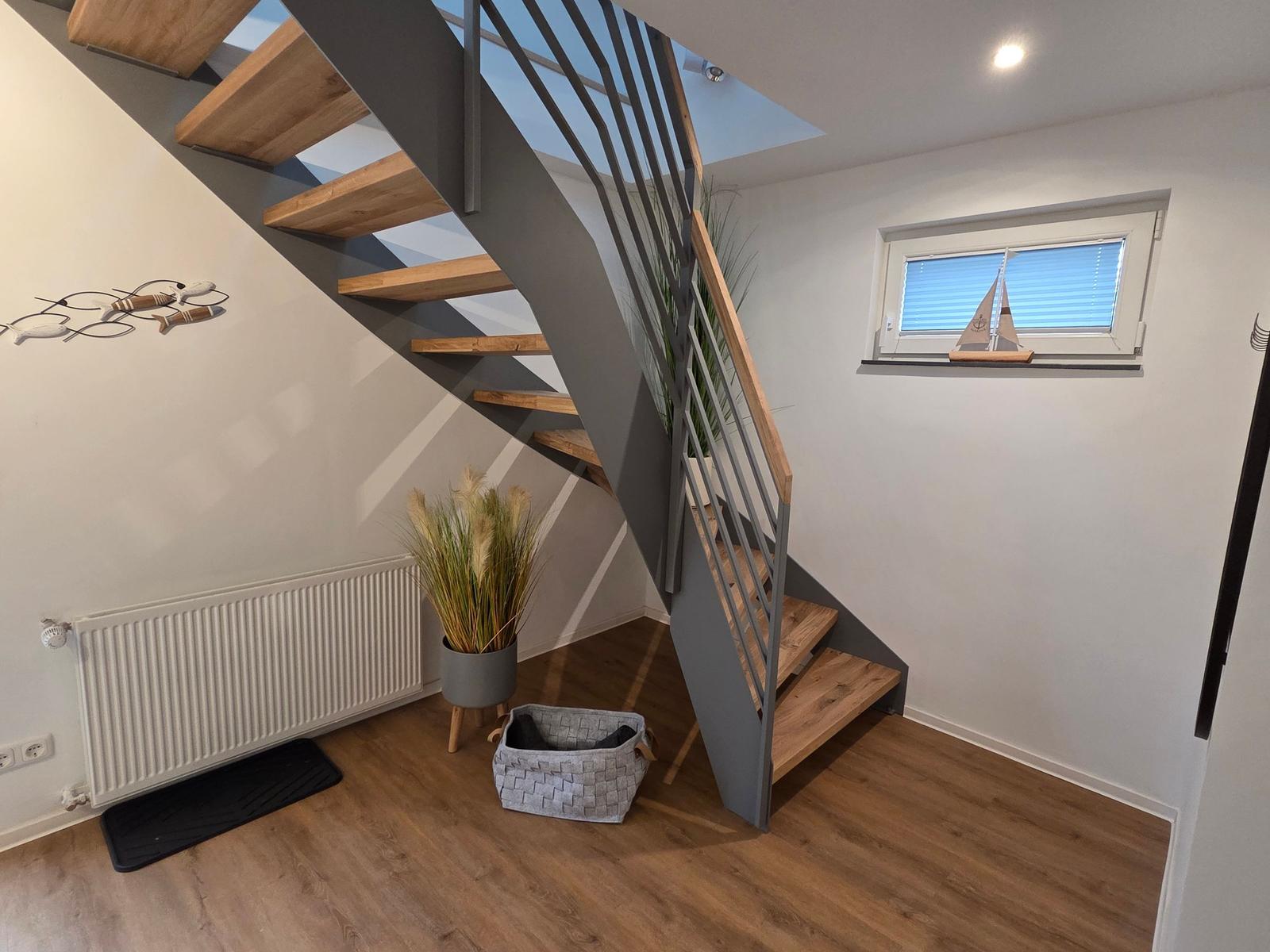 Treppe mit Holzstufen und Metallgeländer. Fenster mit Segelschiffmodell. Pflanze und Wärmekörper an der Wand.