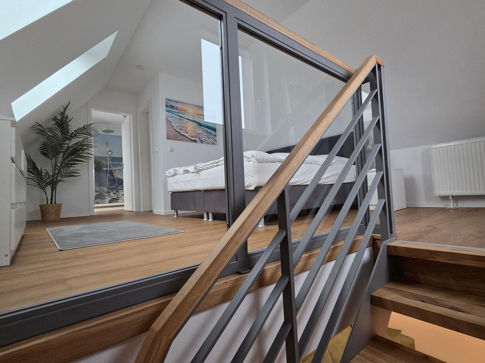 Treppenhaus mit Glasgeländer führt zu einem Schlafzimmer mit Dachschrägen und Holzboden.