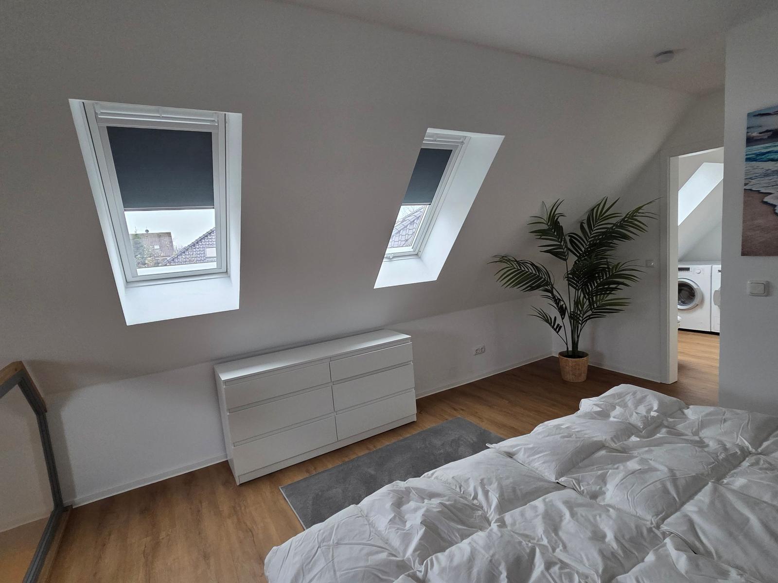Schlafzimmer mit Dachfenstern, Bett, Schrank und Zimmerpflanze. Durchgang zur Waschküche.