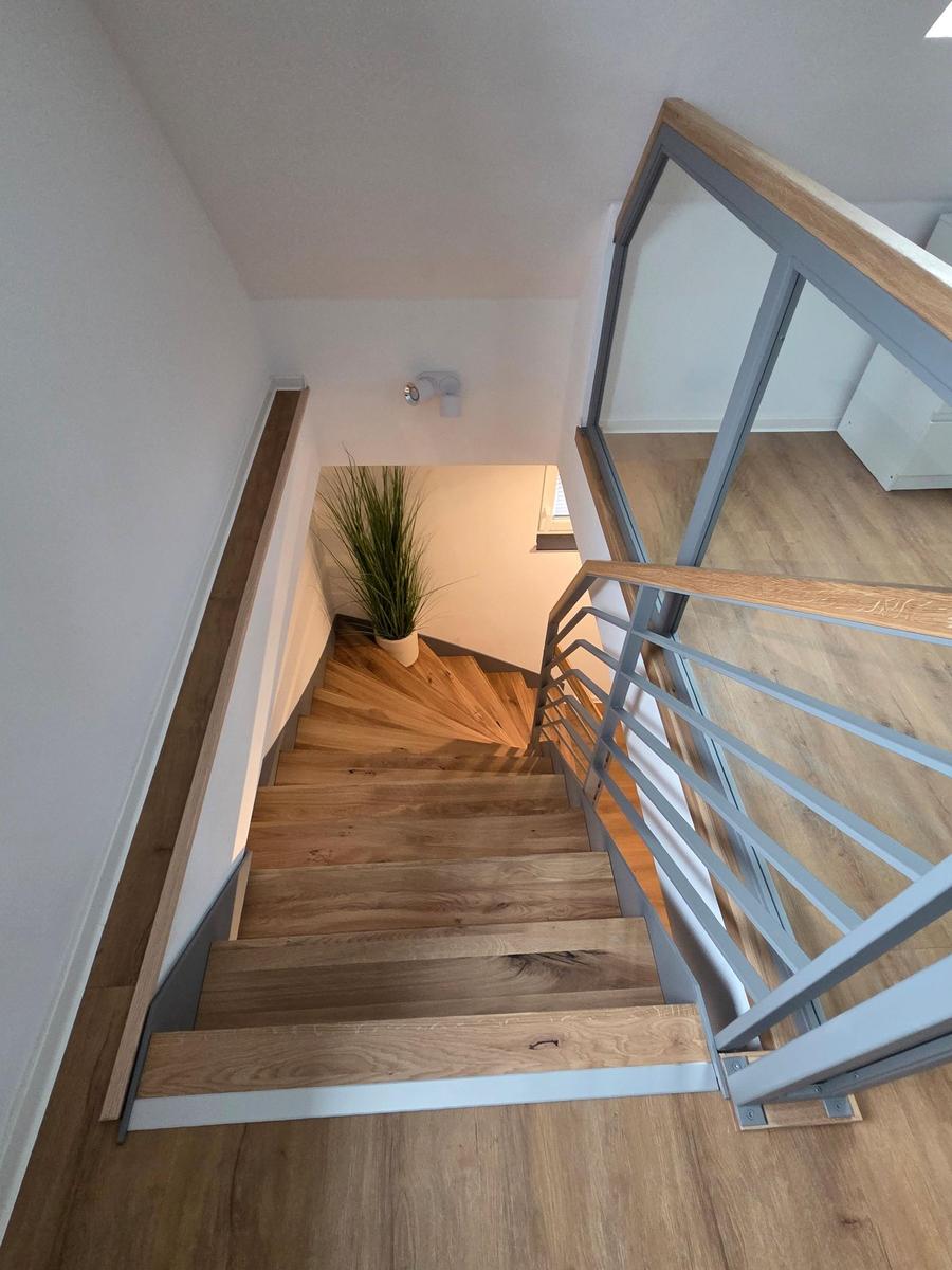 Holztreppe mit Metallgeländer führt zur oberen Etage. Pflanze und Licht an der Wand.