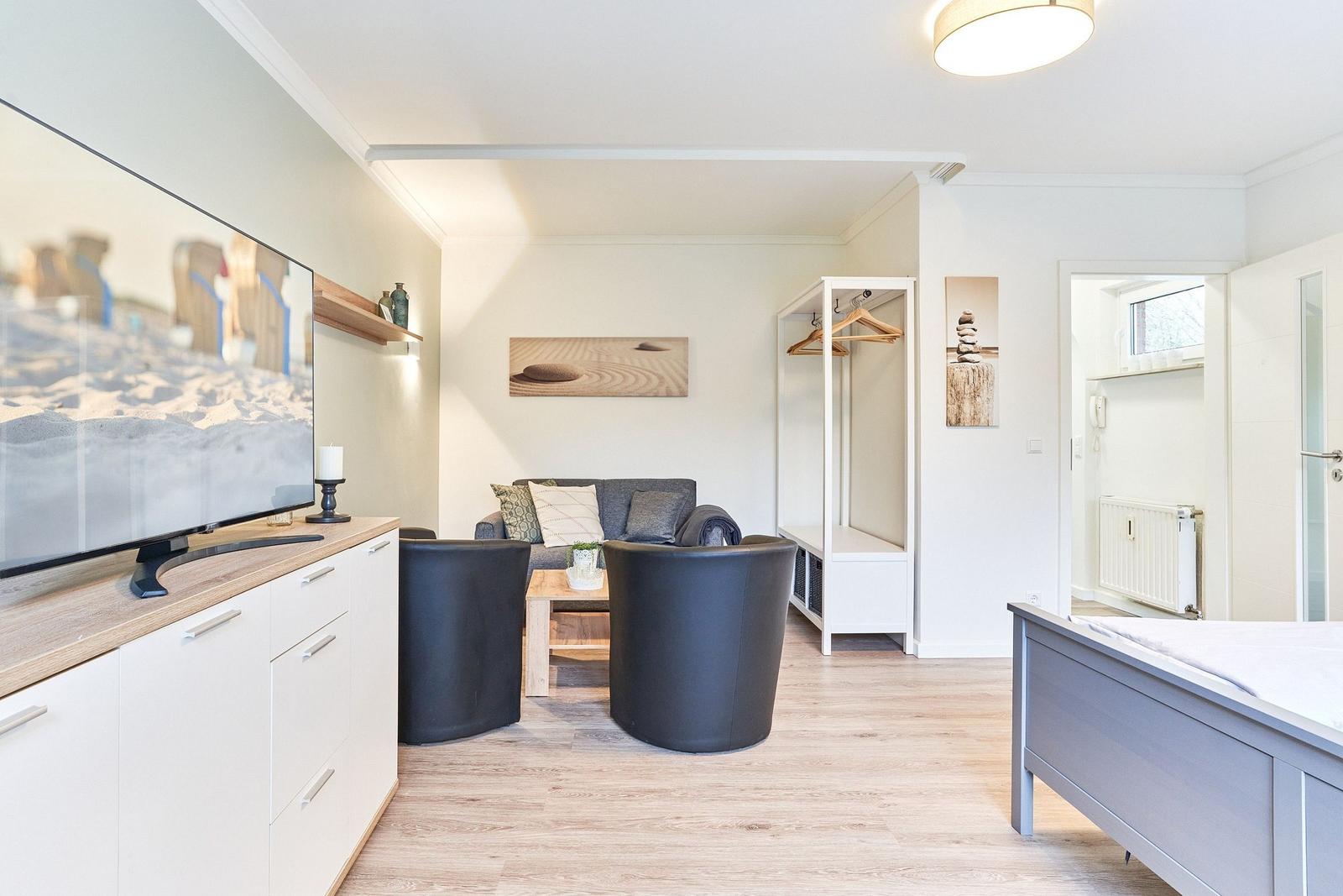 Wohnzimmer mit Sofa, zwei Sesseln und Fernseher. Anschlussraum mit Bett und Kleiderschrank.