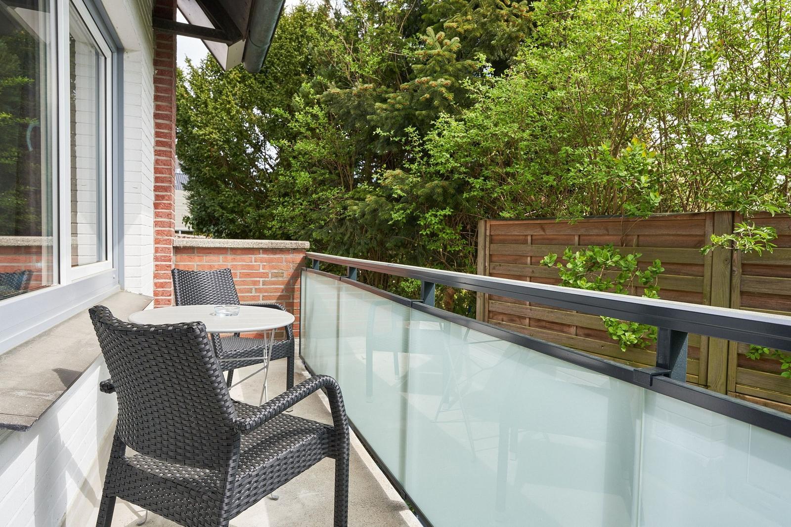 Terrasse mit Tisch und zwei Stühlen, Glasgeländer und grüner Hintergrund.