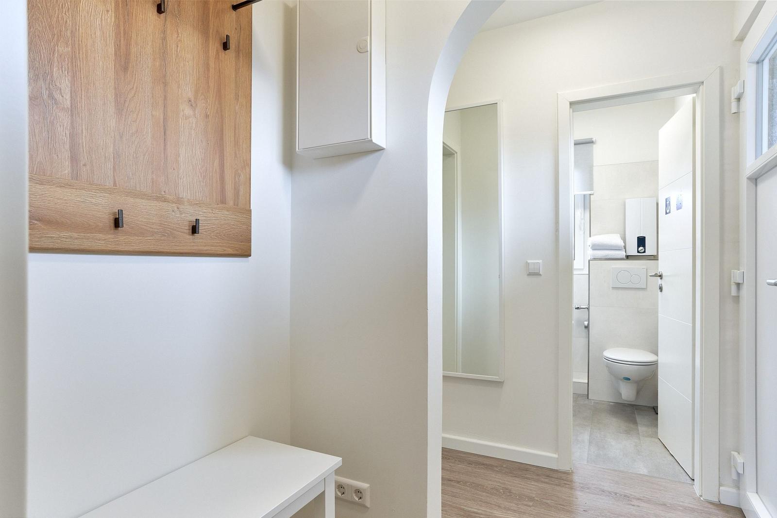 Flur mit Holzregal und weißen Möbeln, Blick in das Badezimmer mit Toilette.