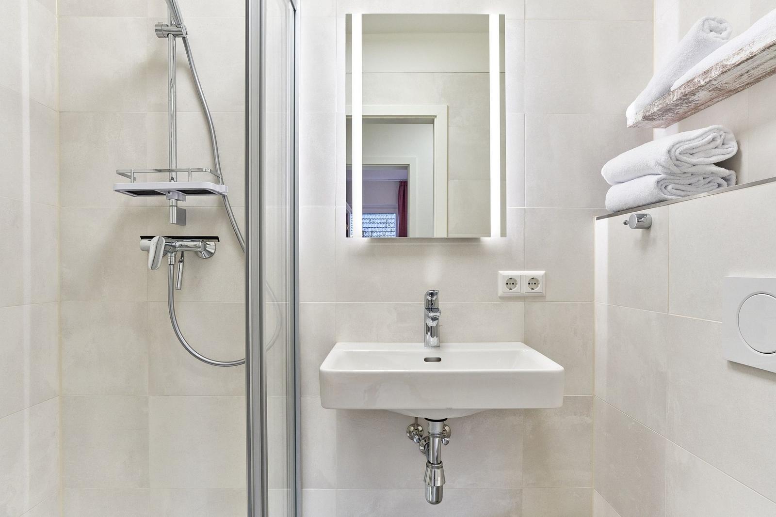 Badezimmer mit Dusche, Waschbecken und Spiegel mit Beleuchtung. Handtücher auf Regal.