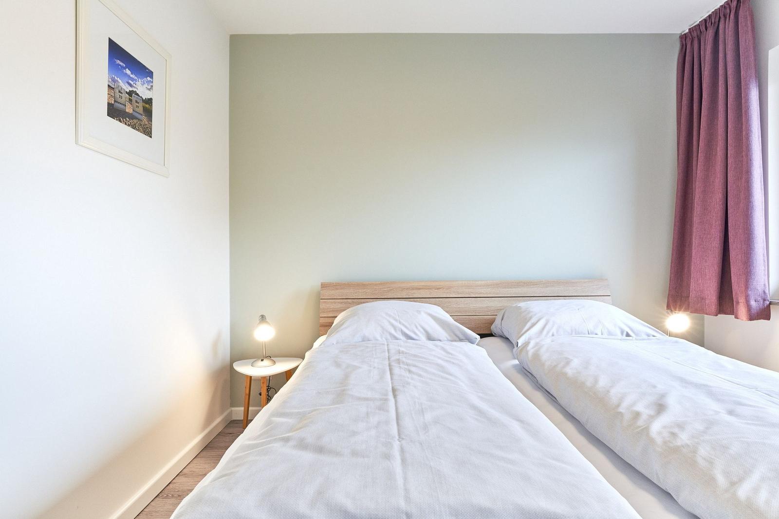 Zwei Einzelbetten mit weißen Bettwäsche, Nachttisch mit Lampe und lila Vorhänge.