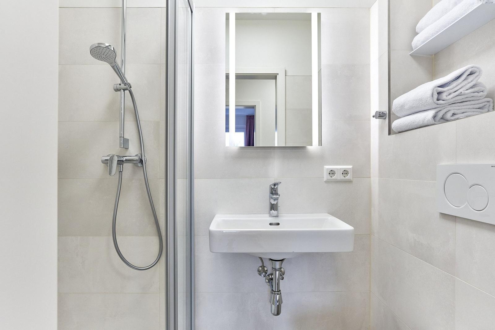 Badezimmer mit Dusche, Waschbecken und Spiegel mit Beleuchtung. Handtücher auf Regal.