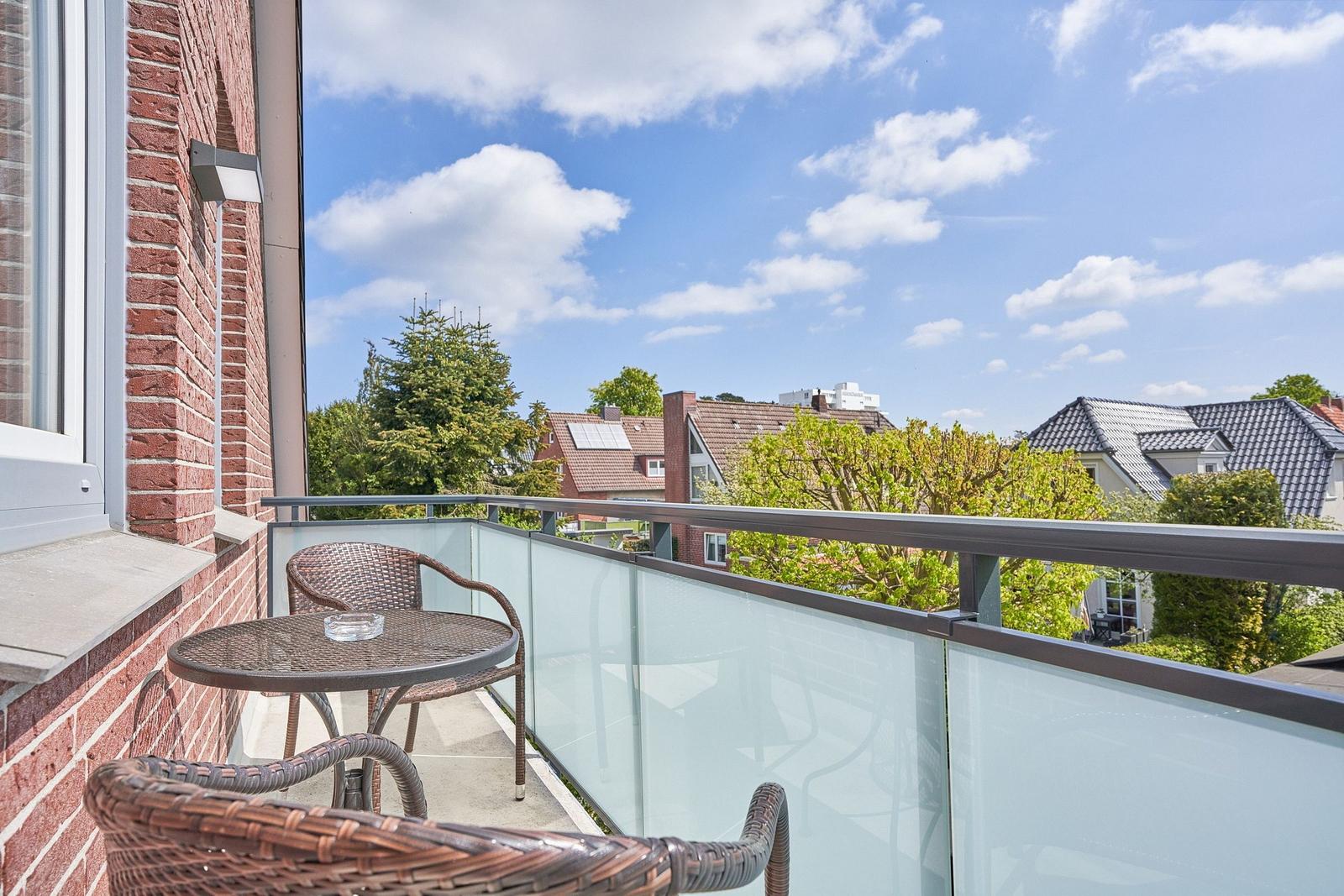 Balcony with wicker furniture and glass railing overlooking rooftops and trees.