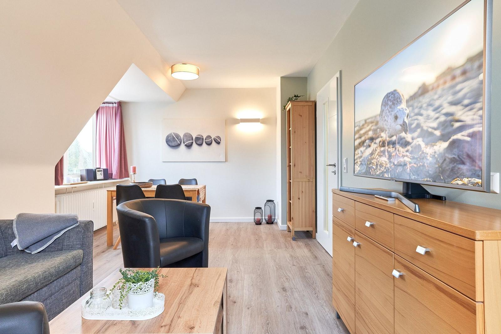 Wohnzimmer mit TV, Sofa, Sessel und Essbereich. Holzboden und große Fenster.