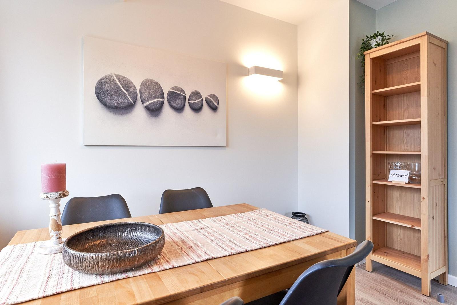 Esszimmer mit Holztisch, Stühlen und Wandbild mit Steinen. Bücherregal mit Pflanze und Glas.