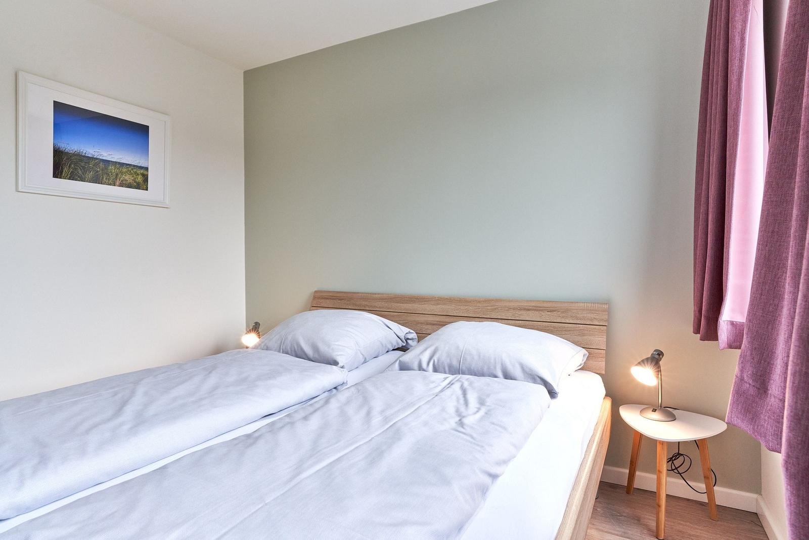 Ein Schlafzimmer mit Doppelbett, Nachttisch und Lampe. Wandbild und lila Vorhänge.