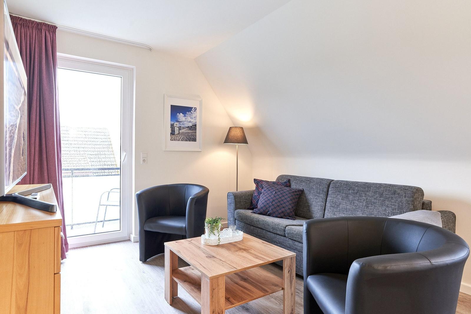 Wohnzimmer mit Sofa, zwei Sesseln, Couchtisch und TV. Fenster zum Balkon.