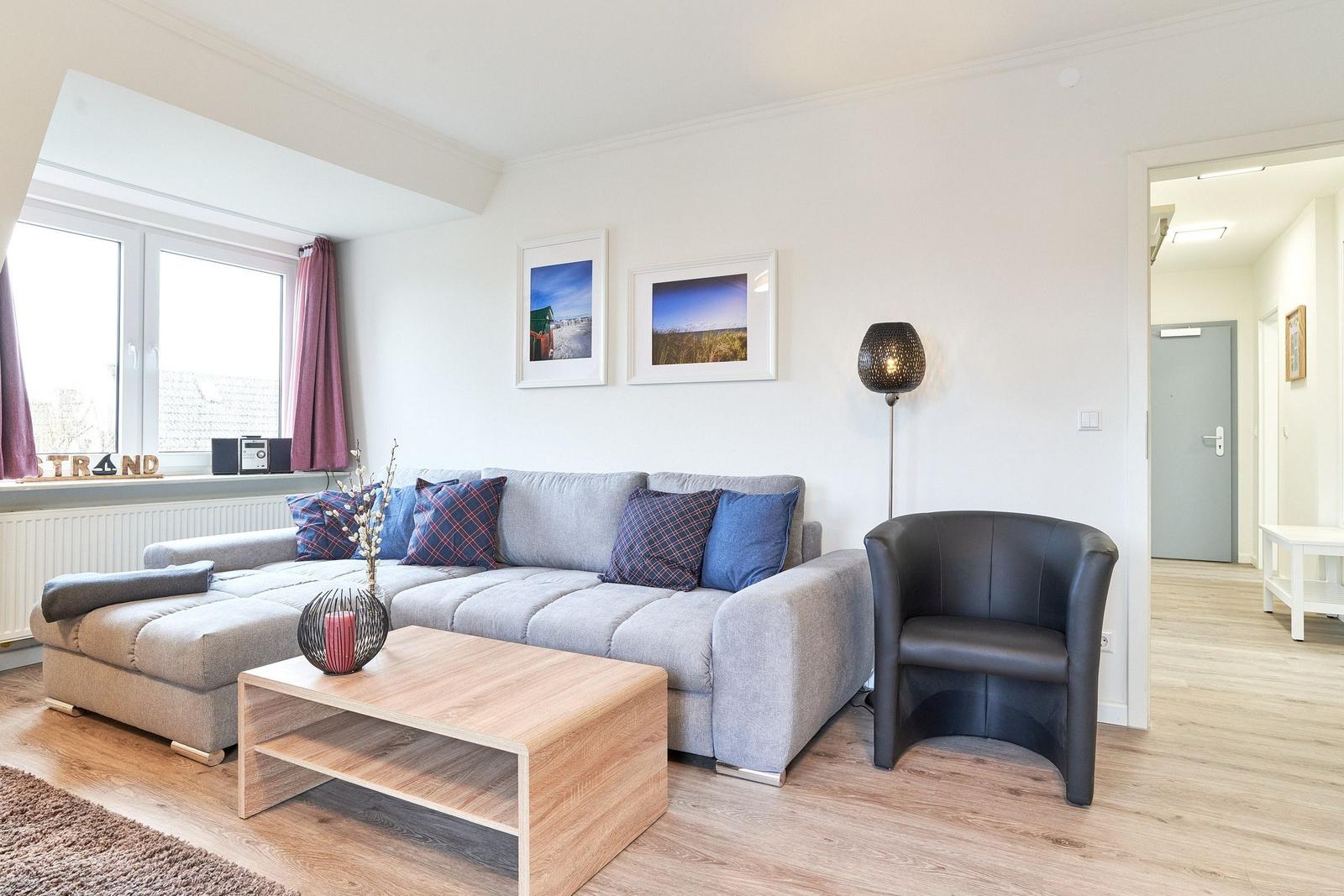 Wohnzimmer mit grauem Sofa, Holz-Tisch und schwarzer Sitzgelegenheit. Fenster mit Vorhängen und Wandbilder.