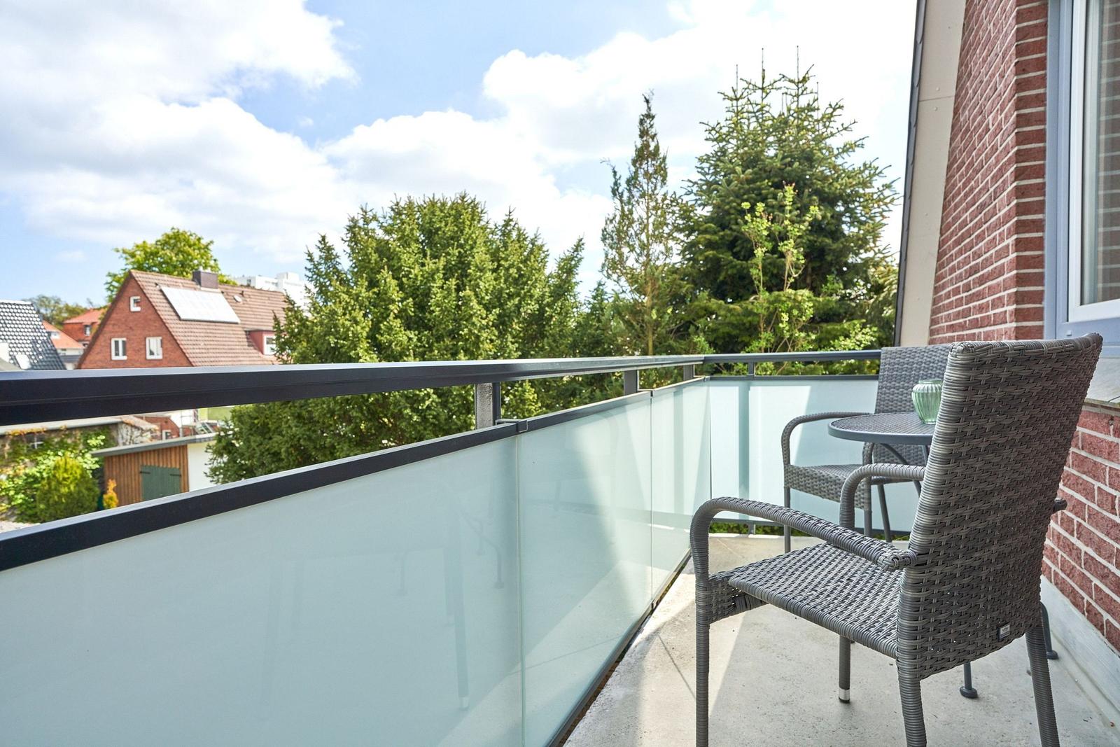 Terrasse mit Glasgeländer und zwei Stühlen. Blick auf grüne Bäume und Gebäude.