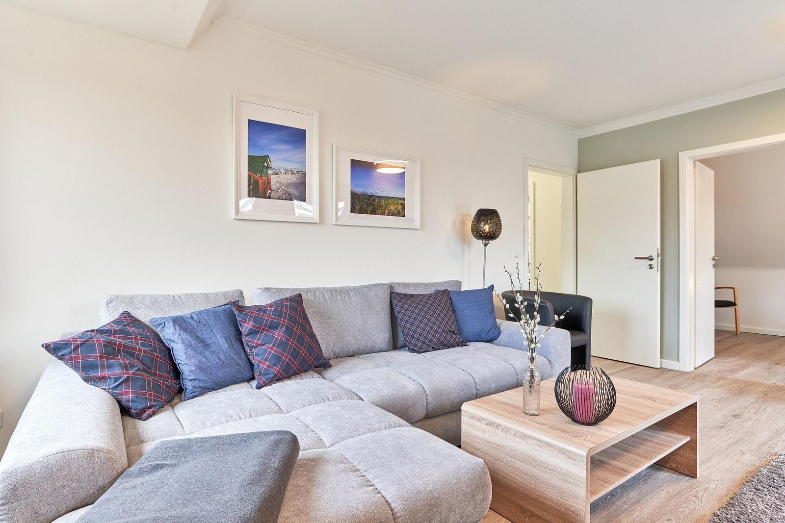 Wohnzimmer mit grauem Sofa, Kissen und Holztisch. Wandbilder und Tür im Hintergrund.