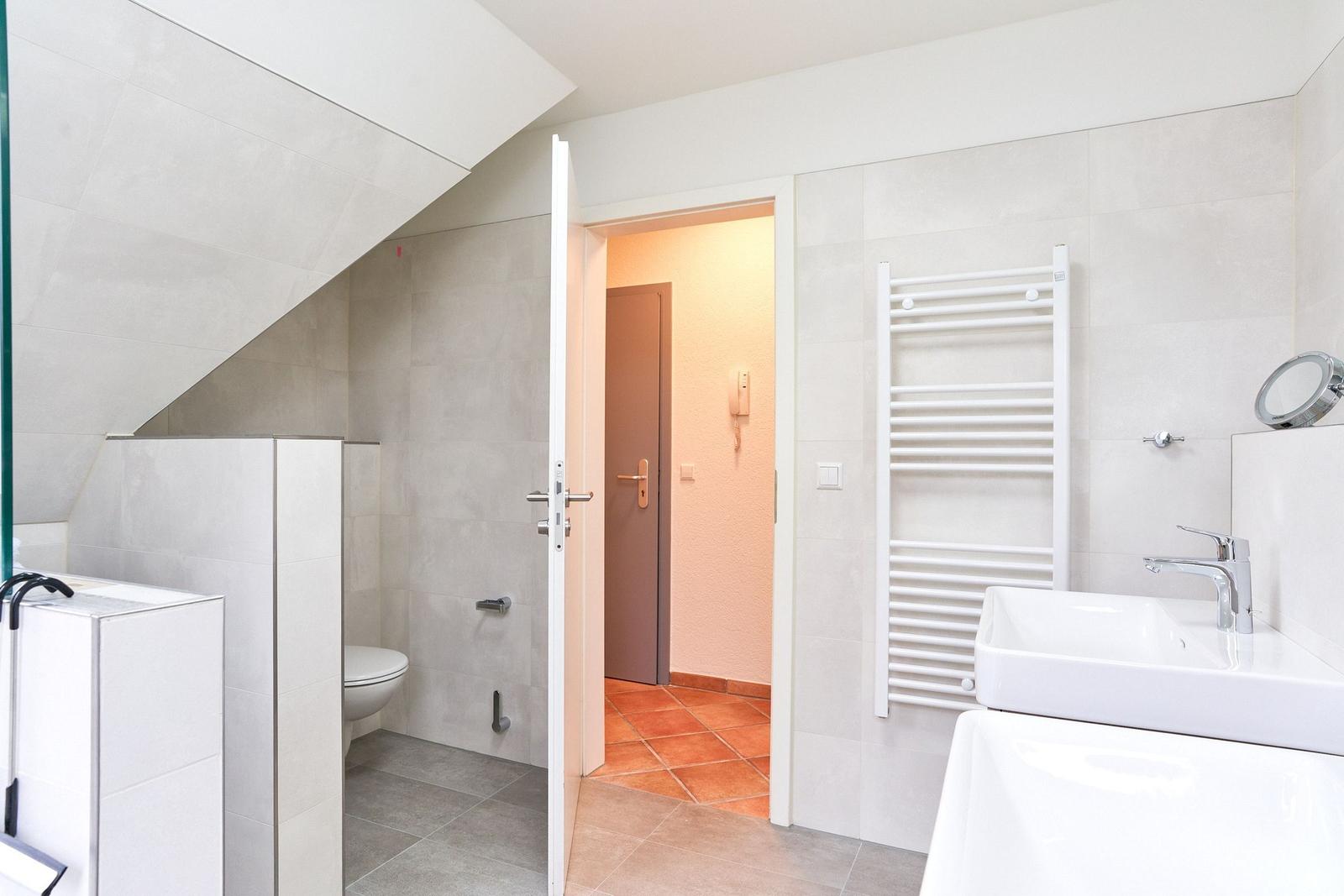 Badezimmer mit Dusche, Waschbecken und Heizkörper. Tür führt in einen Flur.