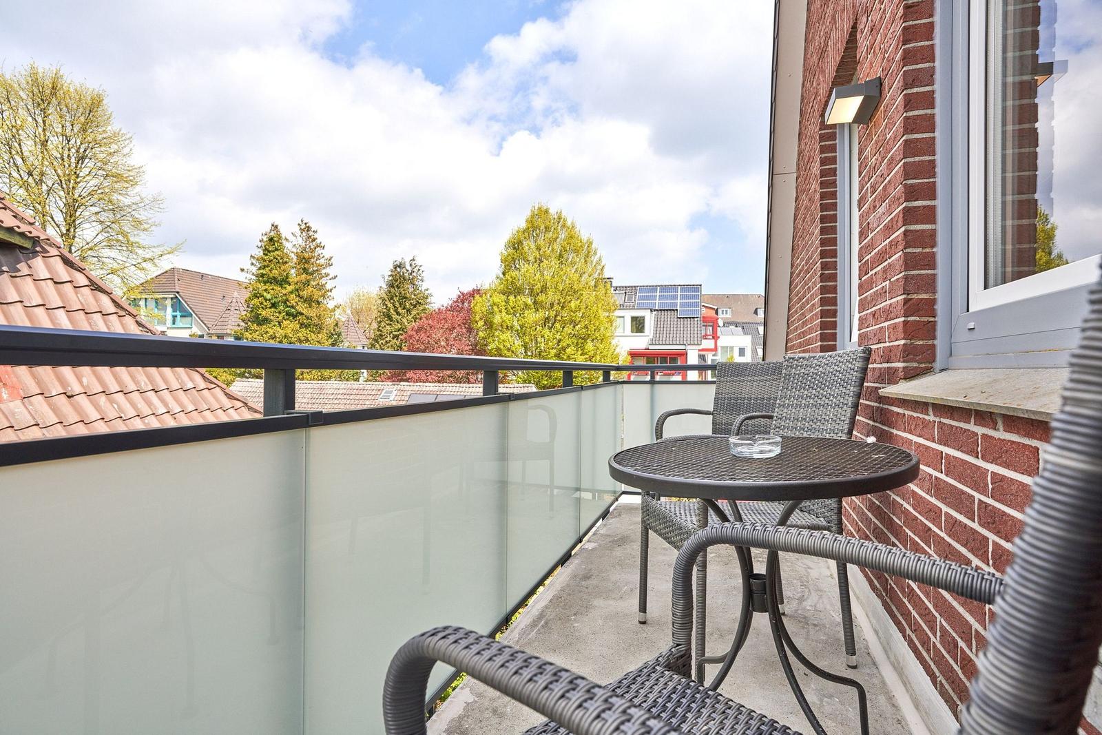 Terrasse mit Tisch und Stühlen, Glasgeländer und Blick auf Bäume und Dächer.