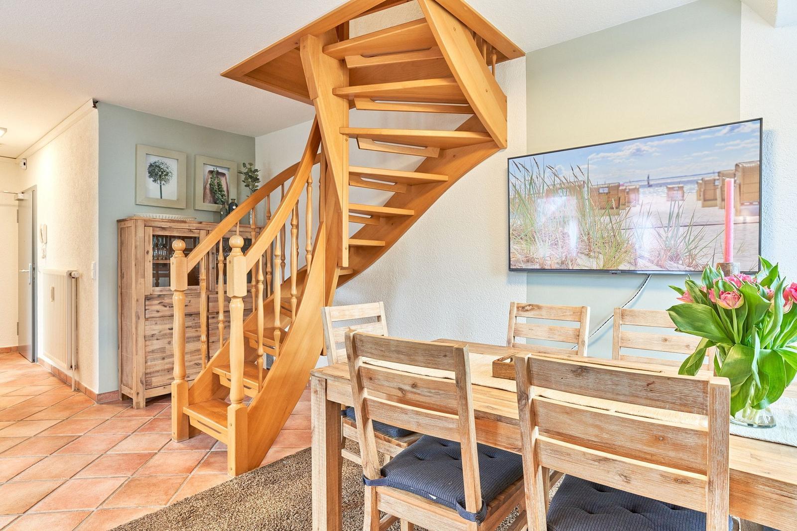 Esszimmer mit Holztisch, Stühlen und spiralförmiger Treppe. Fernseher zeigt Strandbild.