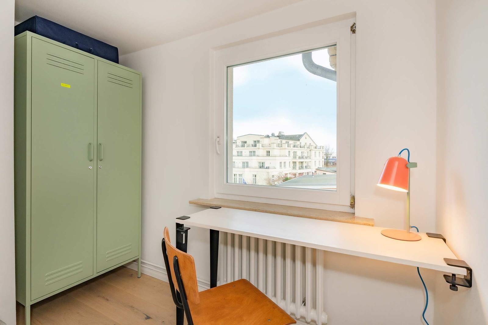 Ein Arbeitszimmer mit Schreibtisch, Stuhl und grünem Schrank. Fensterblick auf Gebäude.