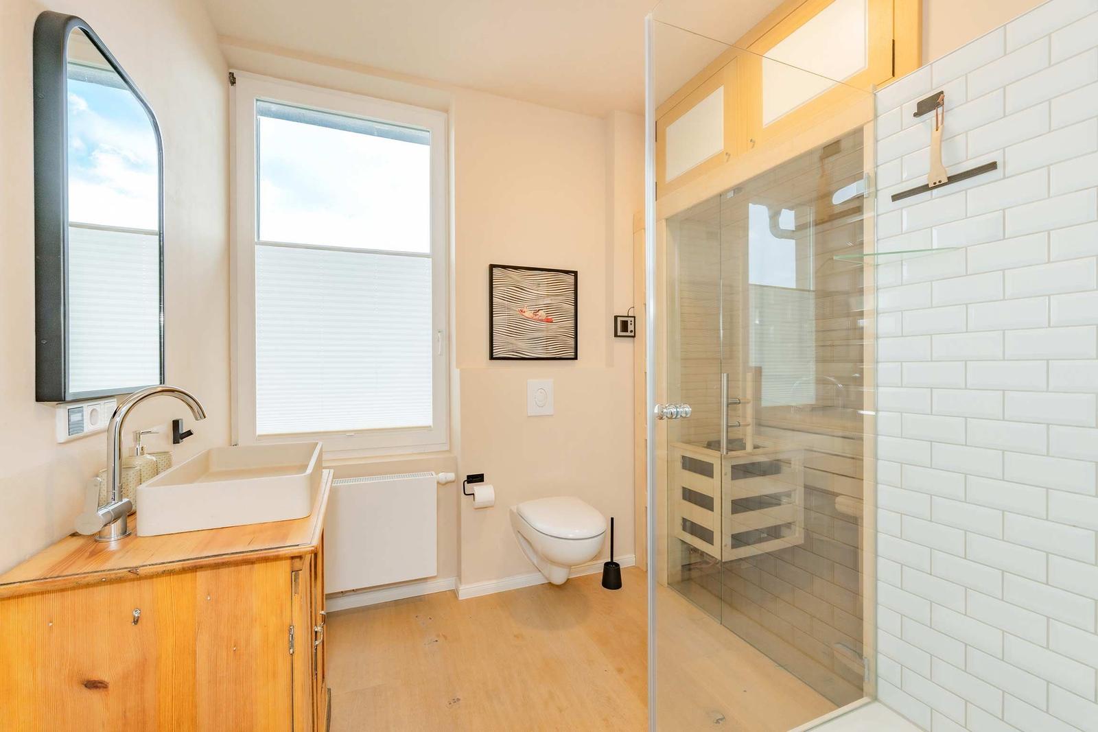 Badezimmer mit Dusche, Waschbecken und Toilette. Holzfußboden und Fenster mit Jalousie.