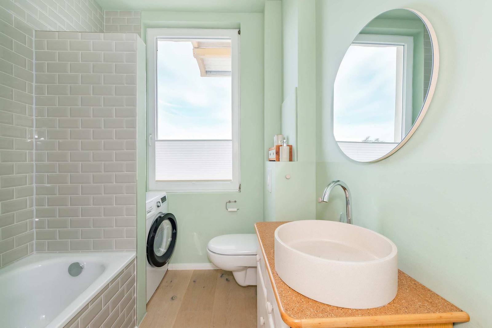 Badezimmer mit Badewanne, Waschmaschine und Waschtisch mit runden Spiegel.