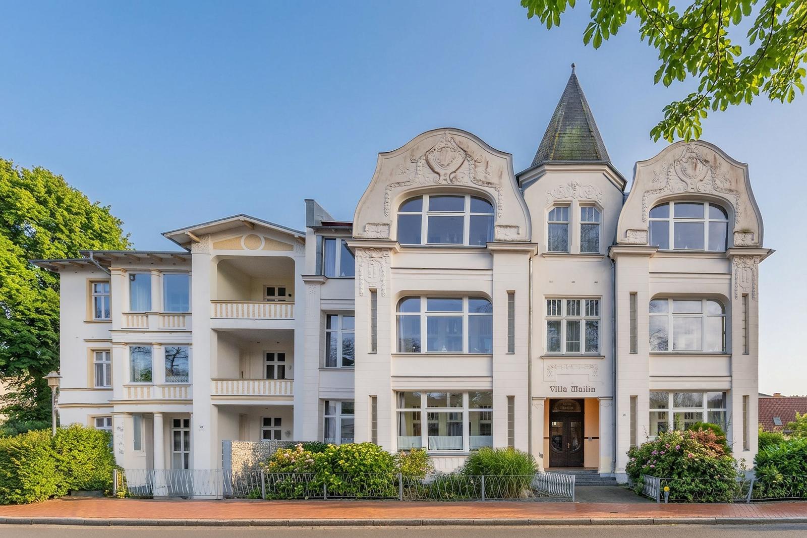 Villa Mailin mit markanter Fassade und Turm. Grüne Bäume und Beet umgeben das Gebäude.