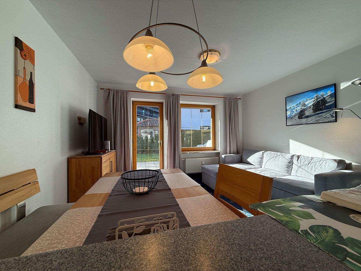 Wohn- und Essbereich mit Sofa, Esstisch und TV. Fenster und Tür zur Außenterrasse.