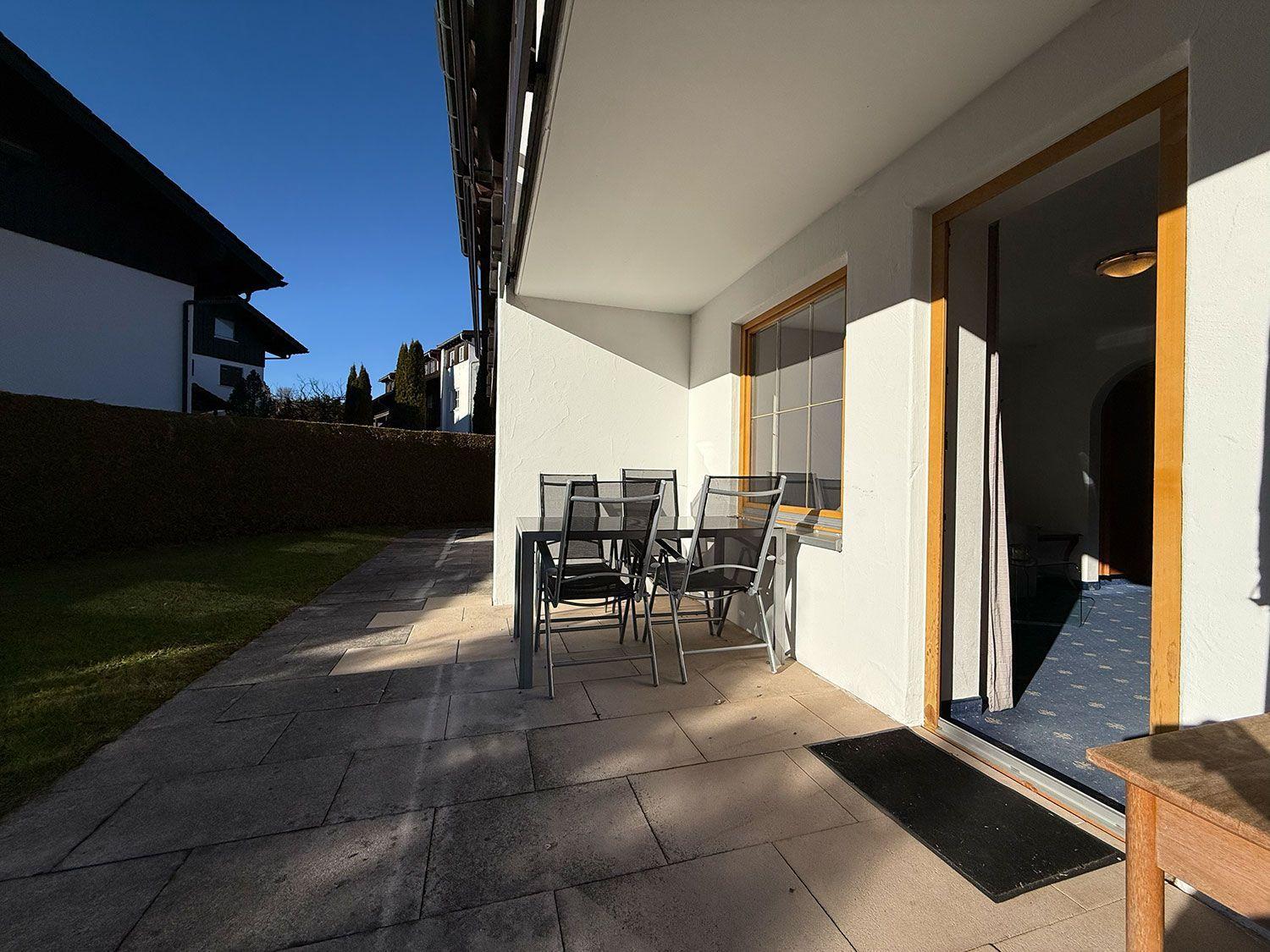 Terrasse mit Tisch und Stühlen, Tür zum Innenraum, Blick auf Garten und Nachbarhaus.