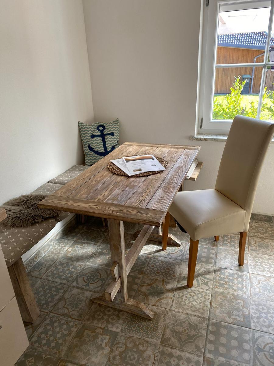 Holztisch mit Stuhl und Sitzbank an Fenster, gemusterte Fliesen und Kissen mit Ankermotiv.