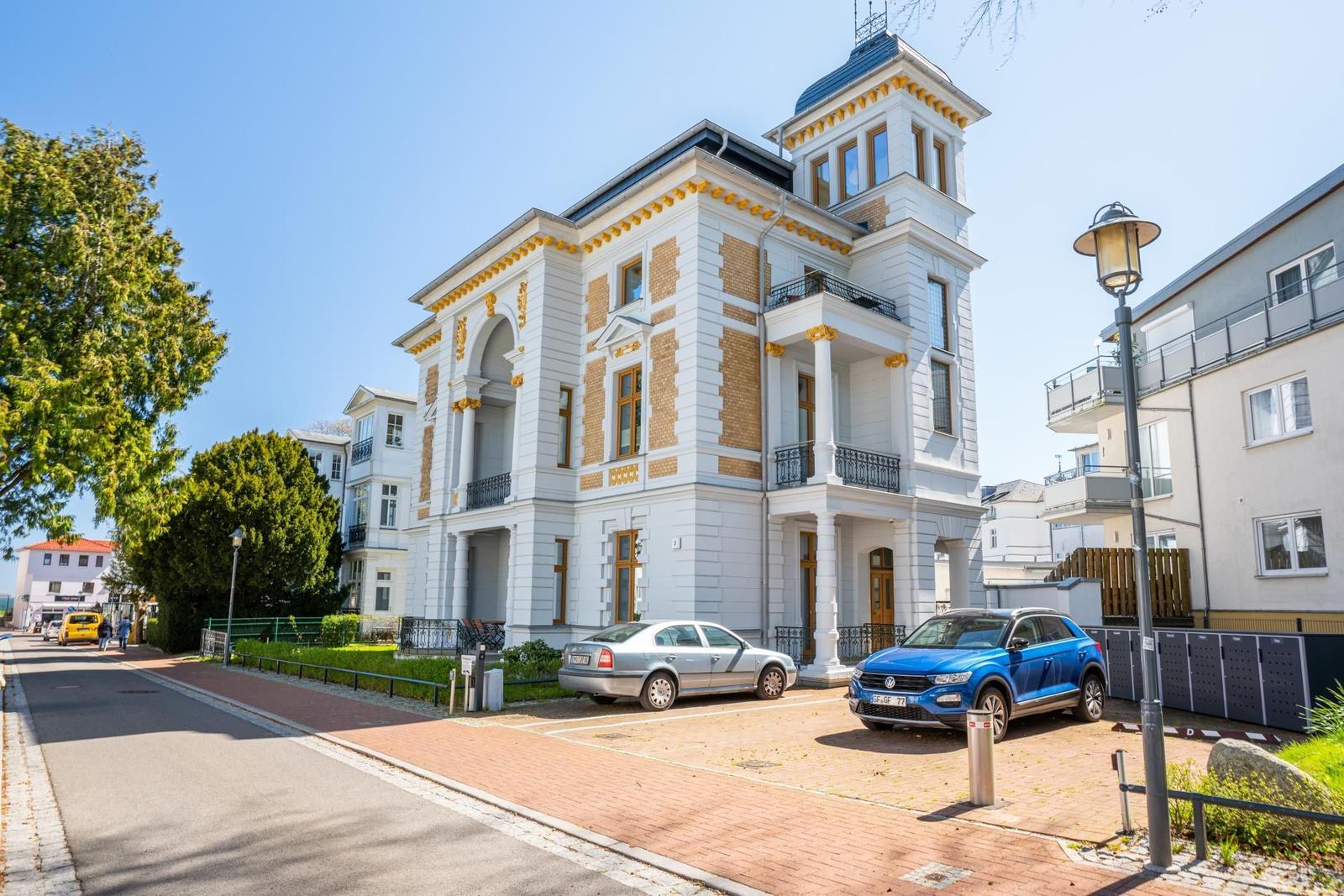 Historisches Gebäude mit weißen Wänden, gelben Akzenten und Balkonen. Zwei Autos parken vor dem Haus.