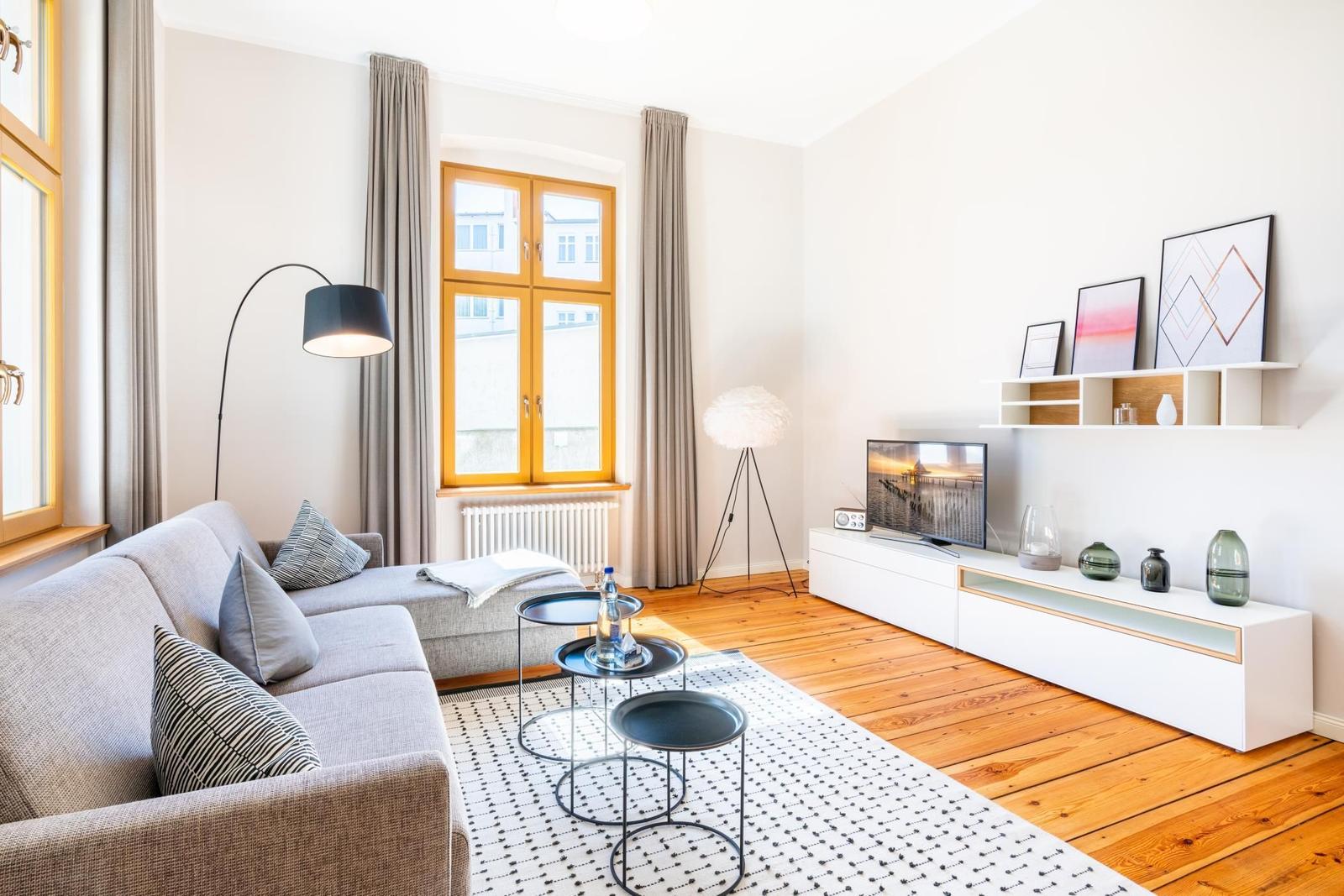 Wohnzimmer mit Sofa, TV und Holzboden. Fenster mit Vorhängen und Beleuchtung.