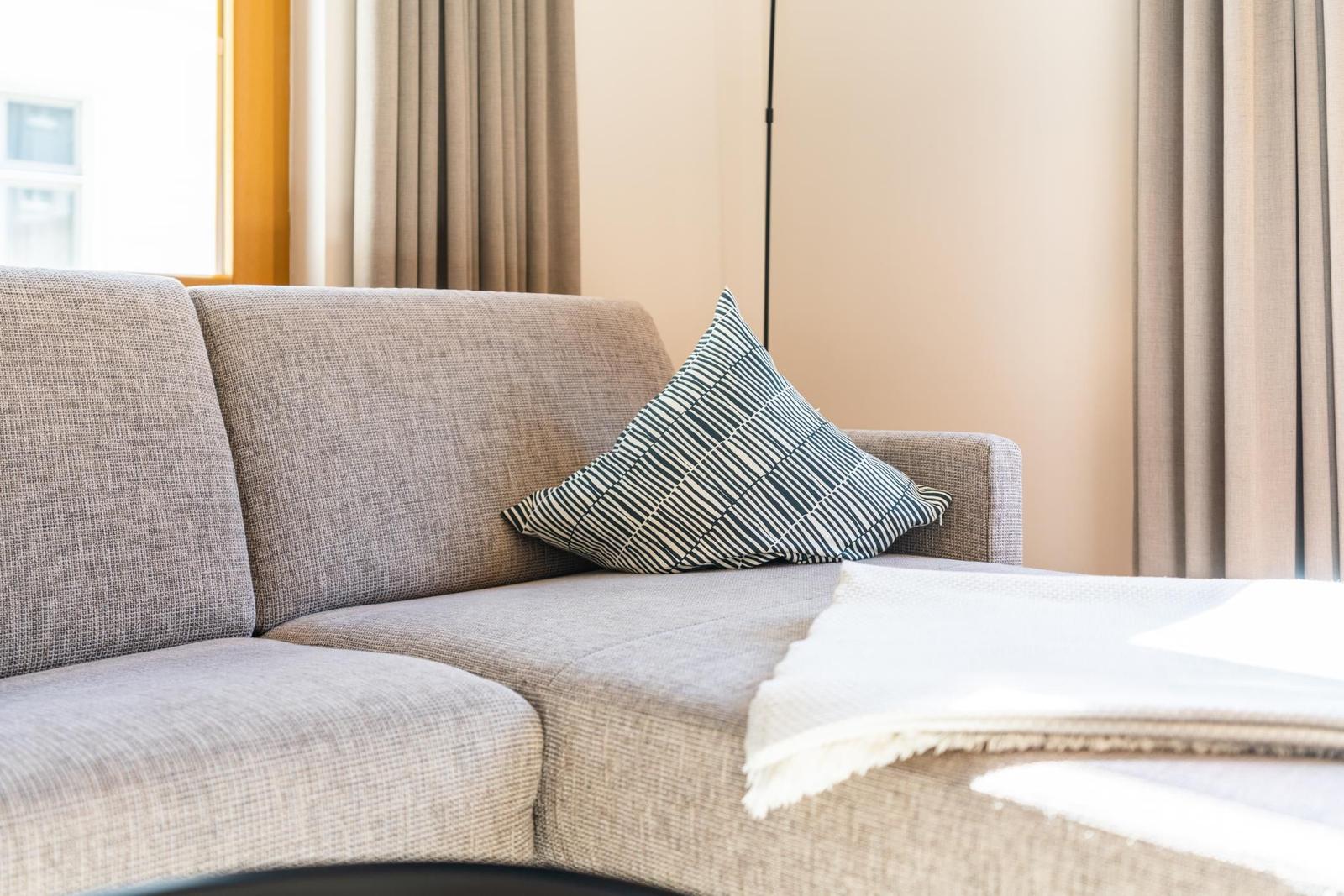 Ein grauer Sofa mit Kissen und Decke vor Fenster mit Vorhängen.